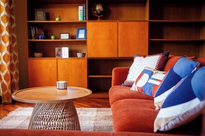 A living room with a round wooden coffee table, a red sofa with patterned cushions, and a bookshelf filled with books and decorative items. - Villa Acqua Blu: Gorgeous villa walking distance to Lake Garda and its beaches 
 (Toscolano Maderno)