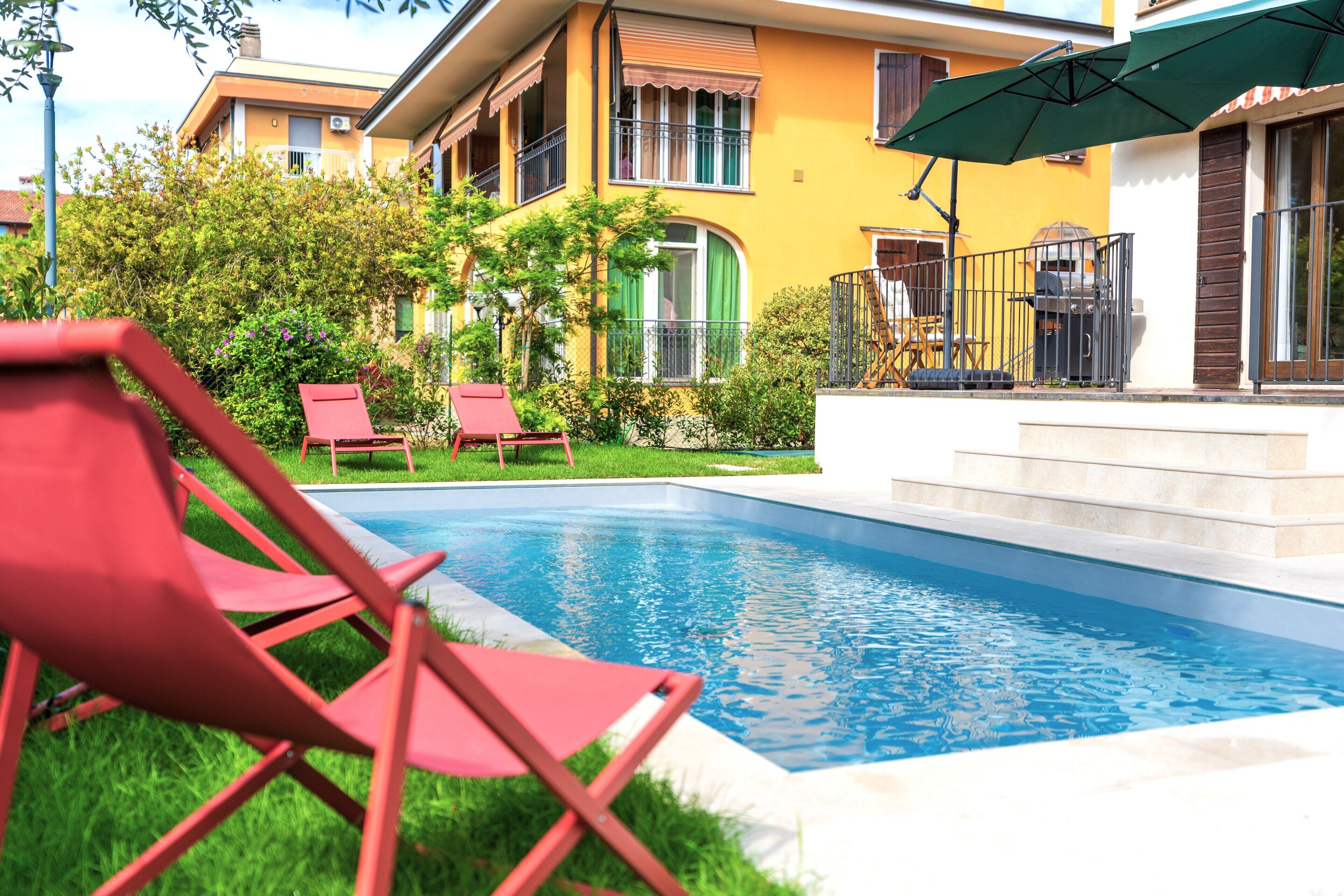 A swimming pool with red lounge chairs, a grill, and a yellow building in the background.