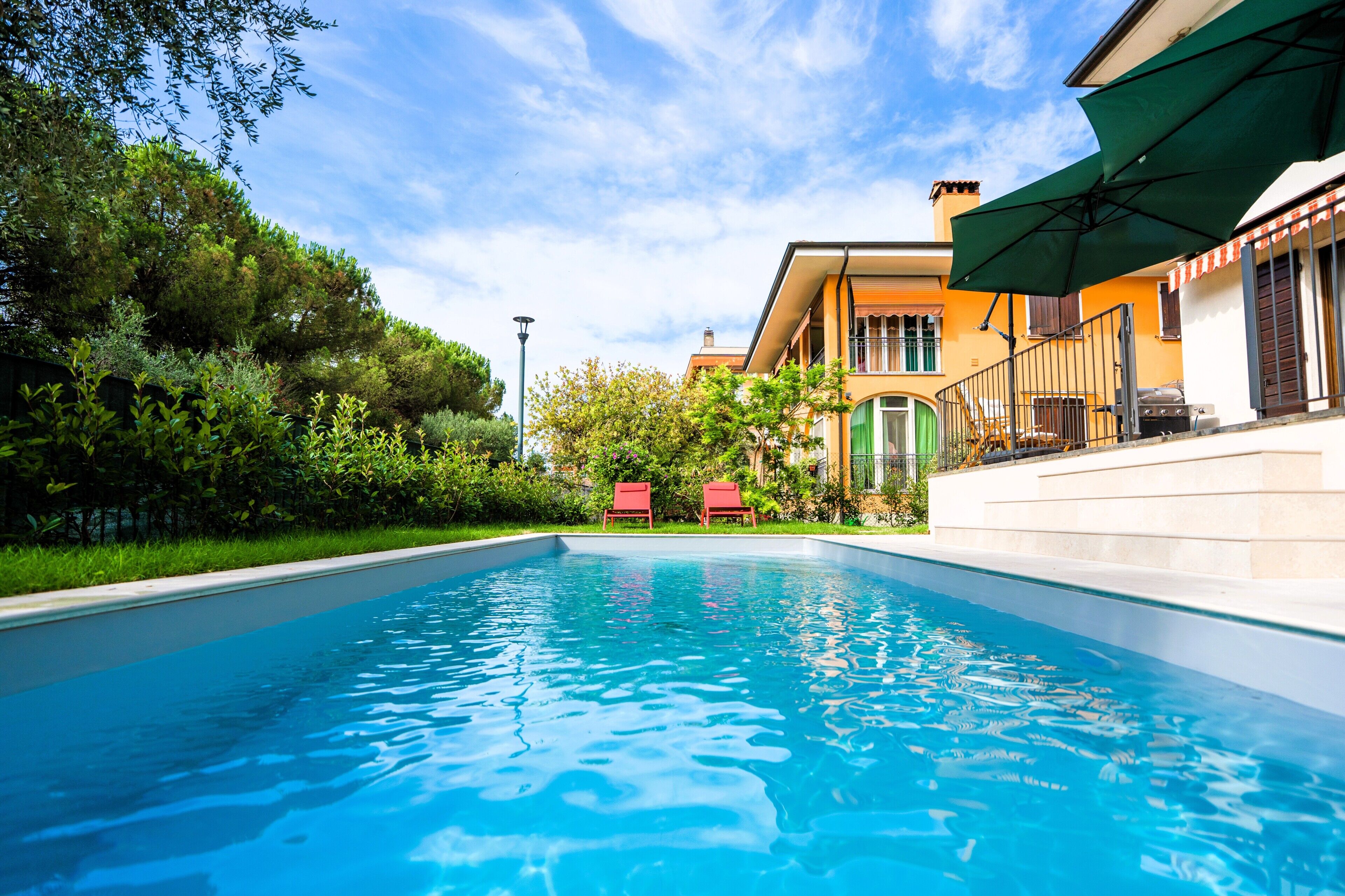 A swimming pool with a clear blue water surface, surrounded by lush greenery and a building with a balcony.