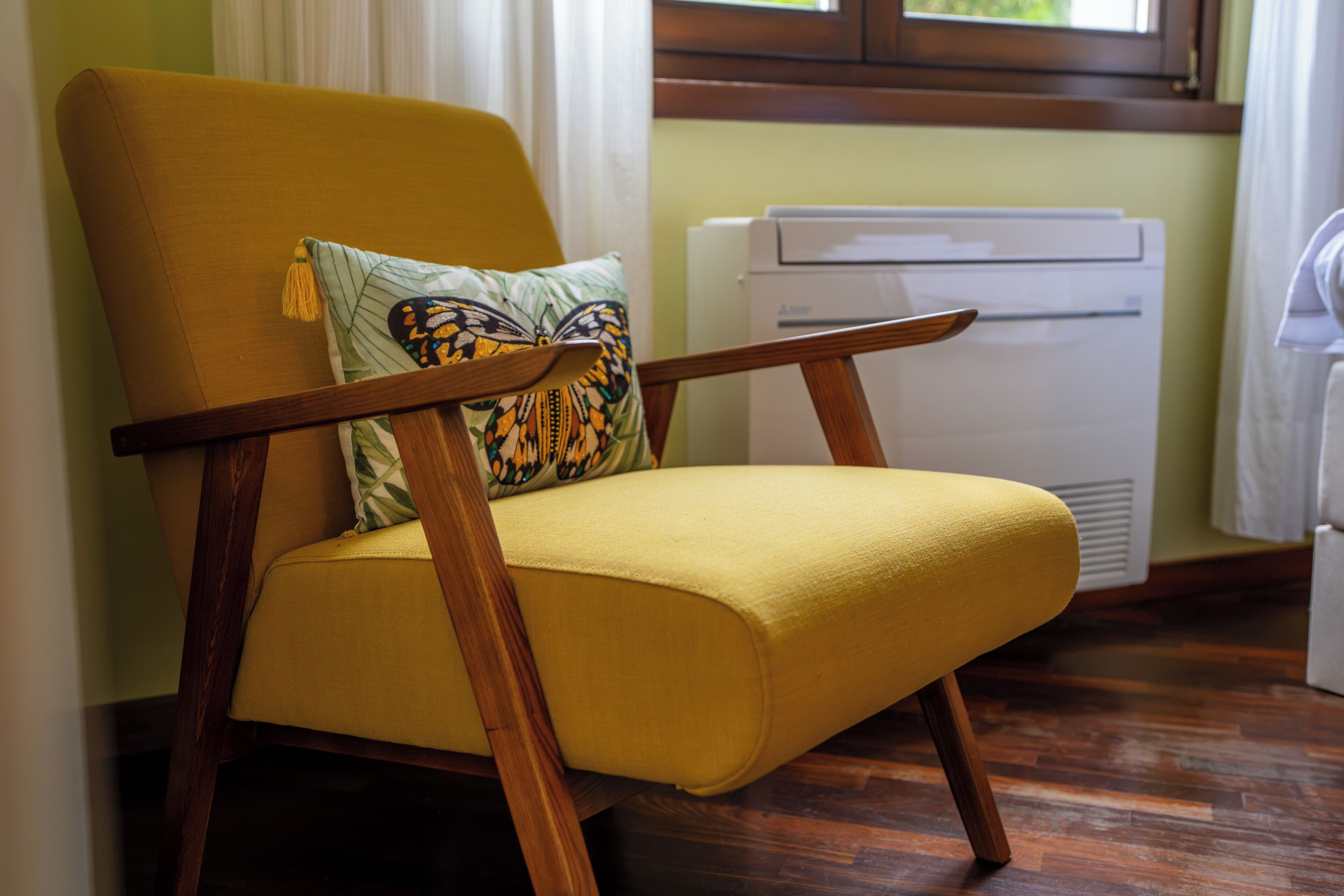 A yellow armchair with a wooden frame and a decorative pillow, positioned in a room with wooden flooring and a white radiator.