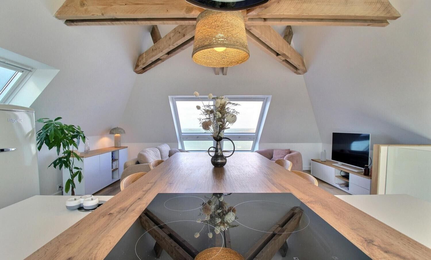 Una habitación moderna estilo loft con una gran mesa de comedor de madera, una lámpara colgante de fibra, un ventanal con vistas, un televisor de pantalla plana y una cocina integrada.