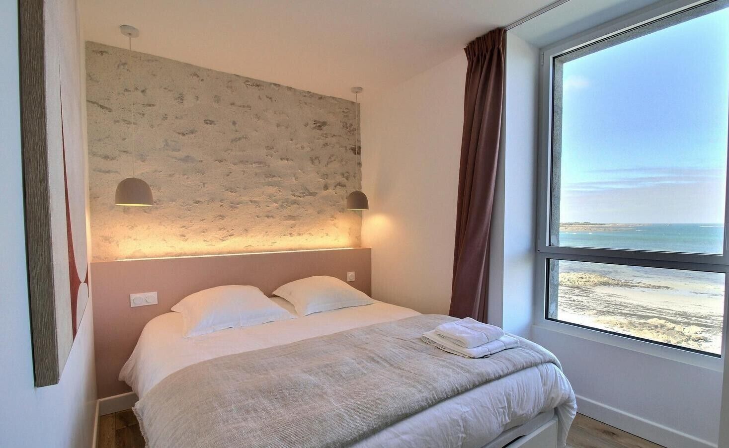 A bedroom with a bed, a headboard with wall art, two wall-mounted lamps, and a large window with a beach view.