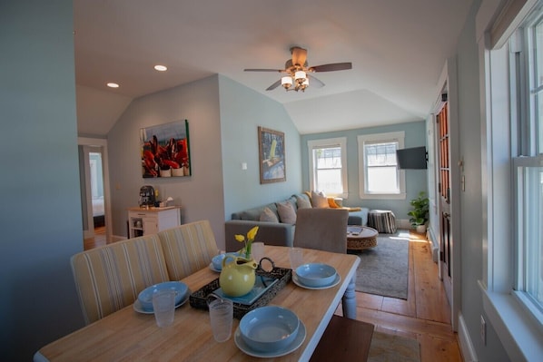 A dining area with a wooden table, chairs, and a ceiling fan. There is a sofa and a window in the background.