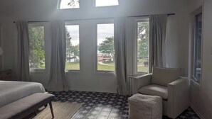 Un dormitorio con cama, una silla, un pequeño reposapiés y ventanas amplias con cortinas.