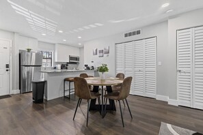 Una cocina moderna con armarios blancos, un refrigerador de acero inoxidable y un microondas. Hay una zona de comedor con una mesa redonda y cuatro sillas.
