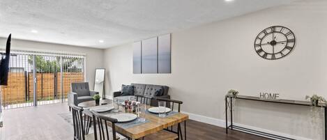 A modern living room with a dining area, a large clock on the wall, and a fireplace with a 'HOME' sign above it.