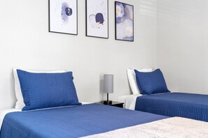 A hotel room with two beds, each with blue bedding and white pillows. There are three framed abstract artworks on the wall above the beds.