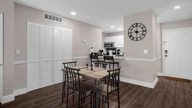 A dining area with a wooden table and chairs, a kitchen in the background, and a clock on the wall.