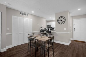 A dining area with a wooden table and chairs, a kitchen in the background, and a clock on the wall.