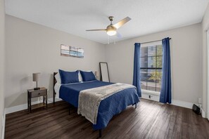 A bedroom with a bed, a nightstand, a ceiling fan, a window with curtains, and a wall art piece.