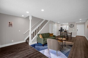 A spacious living room with a green sofa, a beige armchair, and a dining area with a round table. A staircase leads to a kitchen area in the background.