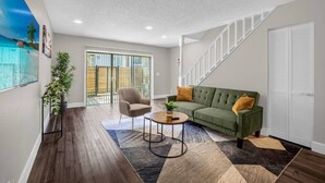 A spacious living room with a green sofa, a coffee table, and a chair. There is a sliding glass door leading to a balcony.