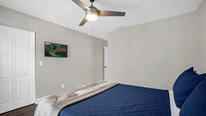A bedroom with a bed, a ceiling fan, a painting on the wall, and a door.