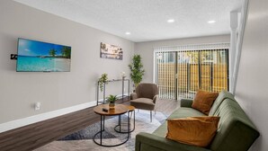 A modern living room with a green sofa, a round coffee table, and a chair. There is a TV mounted on the wall, a painting, and a window with blinds.