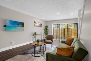 A modern living room with a green sofa, a round coffee table, and a chair. There is a TV mounted on the wall, a painting, and a window with blinds.