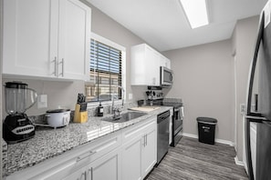 A modern kitchen with white cabinets, a granite countertop, a built-in microwave, a stainless steel refrigerator, and a window with blinds.