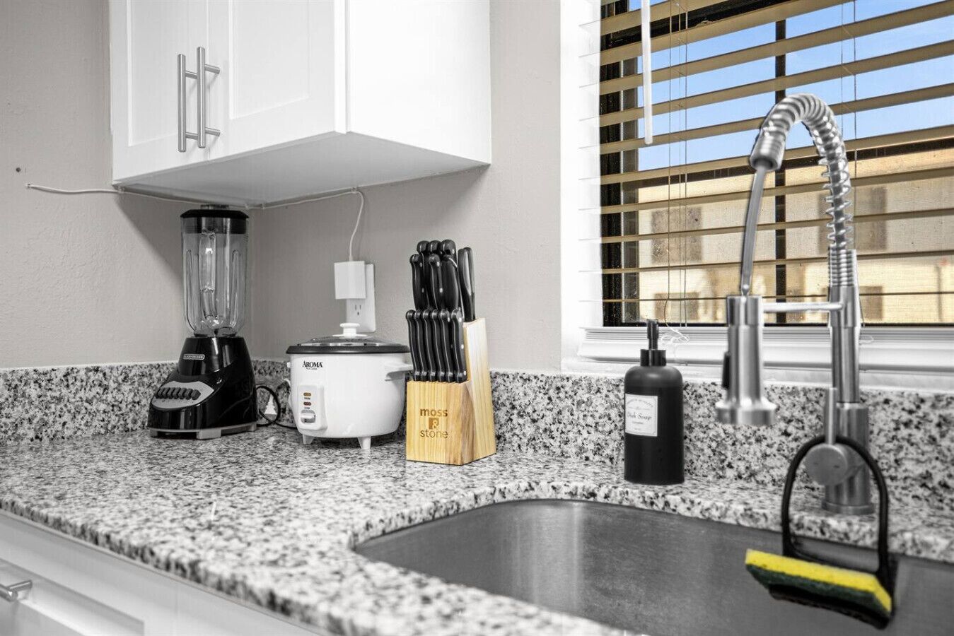 Una cocina con encimera de granito, batidora, olla de cocción lenta, bloque de cuchillos, botella de detergente y pileta de acero inoxidable con grifo.