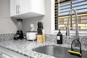 A kitchen with a granite countertop, a blender, a slow cooker, a knife block, a bottle of dish soap, and a stainless steel sink with a faucet.