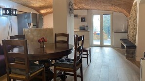 A dining area with a round table and chairs, a kitchen in the background, and a stone wall.