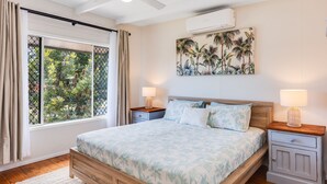 Een slaapkamer met een bed, nachtkastjes, een raam met gordijnen, een airconditioning en een tropisch schilderij.