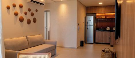 A modern living room with a sofa, a dining table, and a kitchen area with wooden cabinets and a refrigerator.