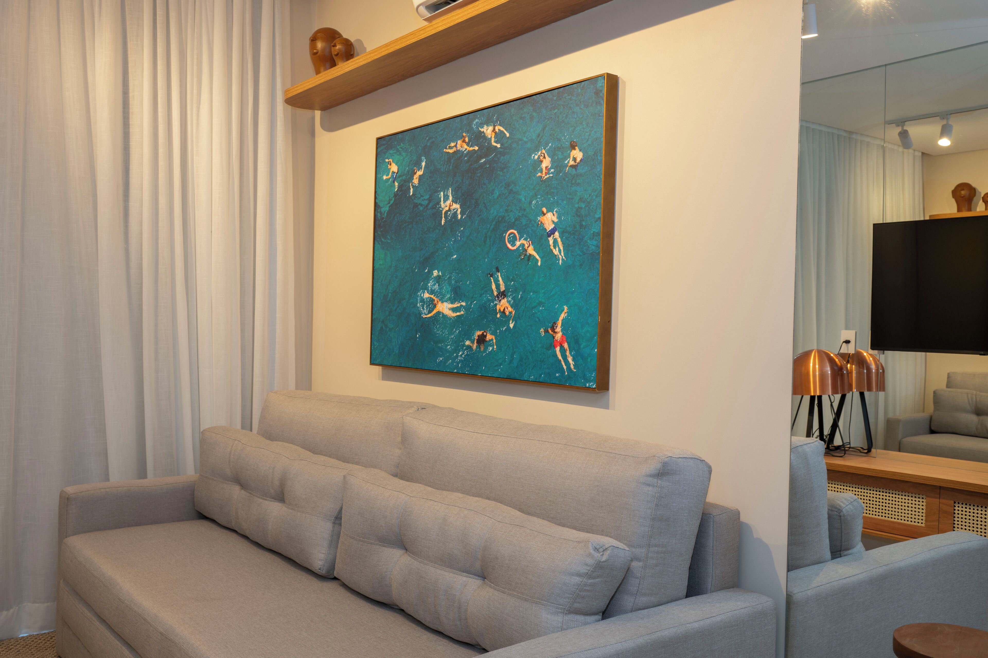 A living room with a grey sofa, a painting of swimmers, and a flat-screen TV on the wall.