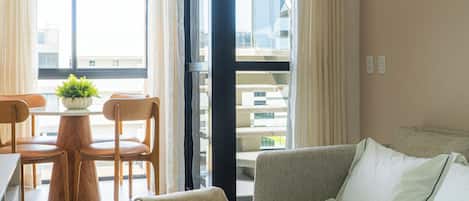 A modern hotel room with a sofa, a dining table with chairs, a potted plant, and a large window with curtains.