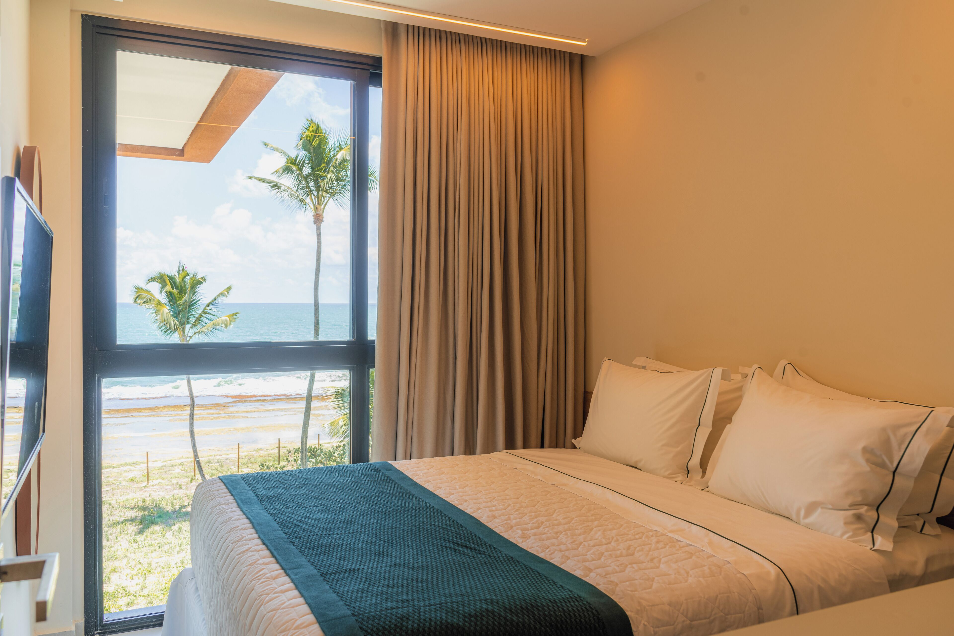 A hotel room with a bed, a large window with a view of the beach and palm trees, and a curtain.