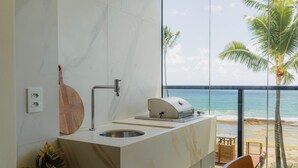 A modern bathroom with a marble vanity, a sink, and a built-in grill. It overlooks a beach with palm trees and the ocean. - Cais Eco Residência Muro Alto by Sintta (Ipojuca)