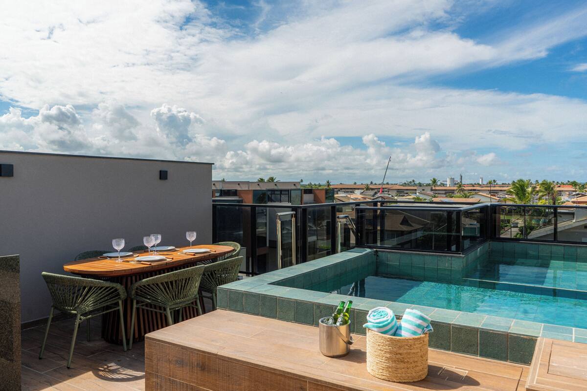 Zona de piscina en la azotea con una mesa de comedor y sillas, una tumbona y una cesta de mimbre con botellas y una toalla.