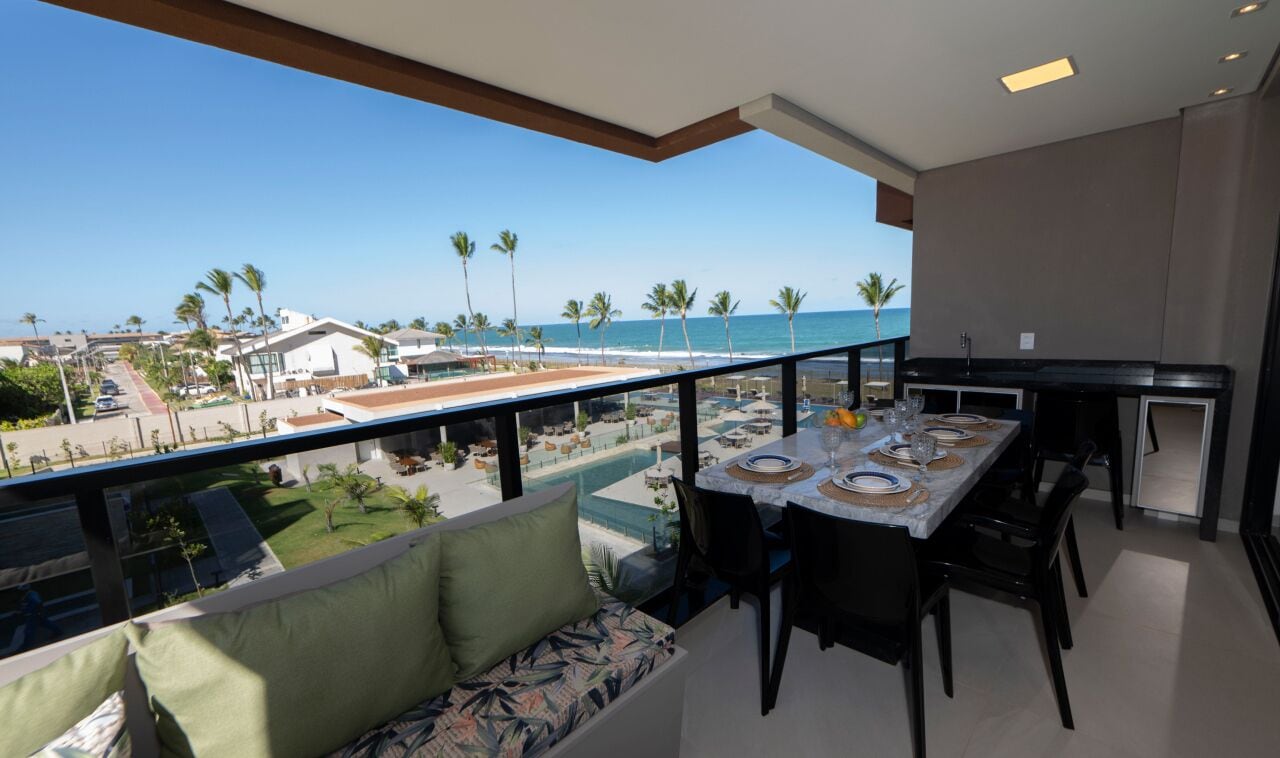 A balcony with a dining table set for four, overlooking a beach with palm trees and a pool.