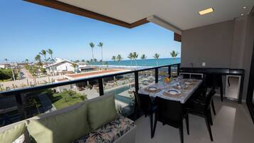 A balcony with a dining table set for four, overlooking a beach with palm trees and a pool.