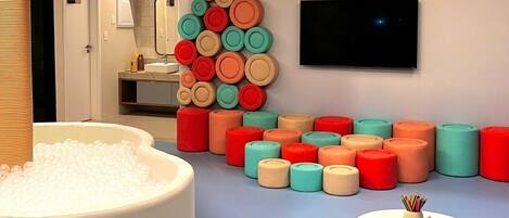 A modern bathroom with a large bathtub filled with bubbles, a wall-mounted TV, and a colorful arrangement of soap bars.