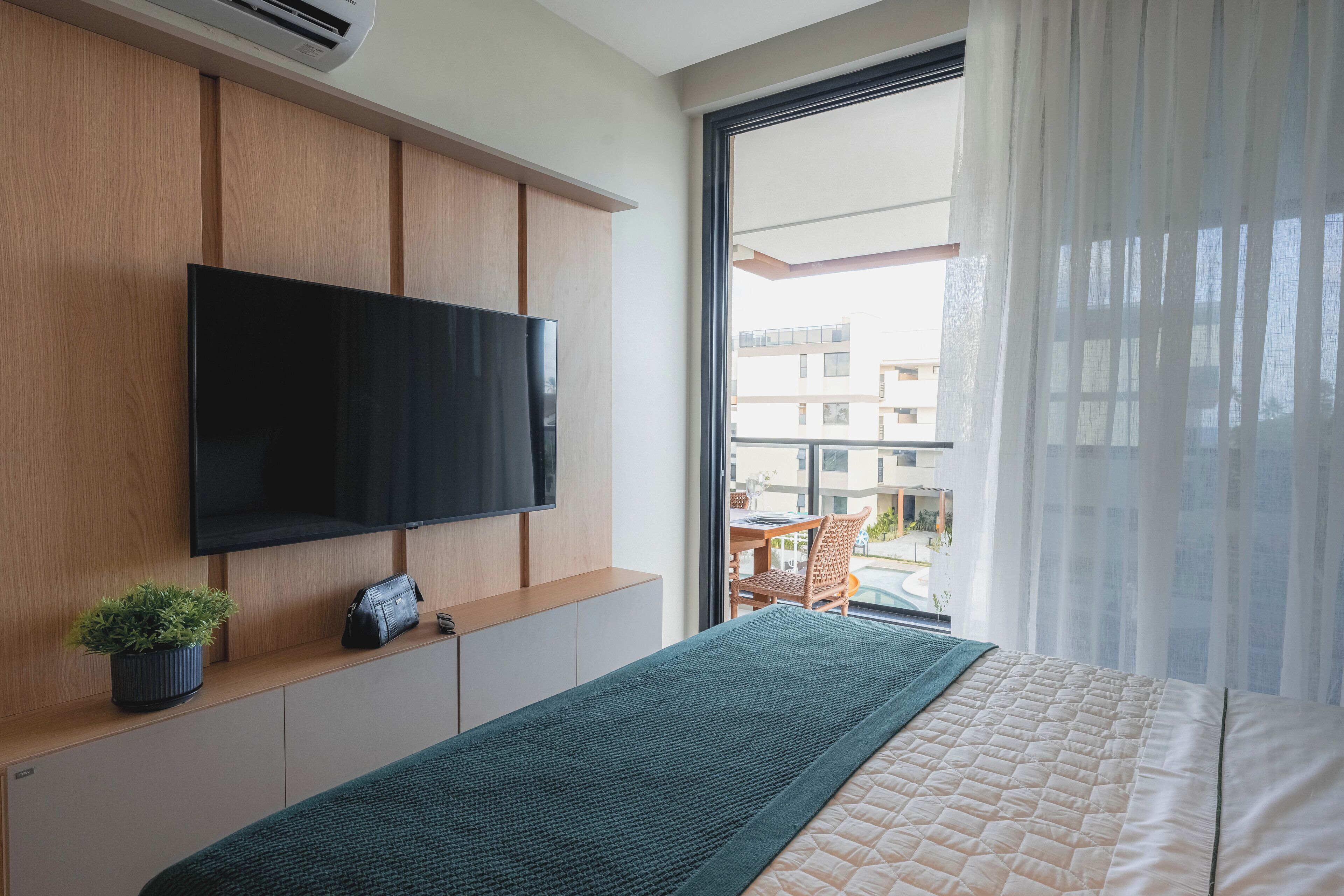 Habitación de hotel moderna con un televisor de pantalla plana grande montado en un panel de madera, cama con colchón acolchado, una pequeña planta en maceta y vistas al balcón exterior a través de cortinas de tela ligera.