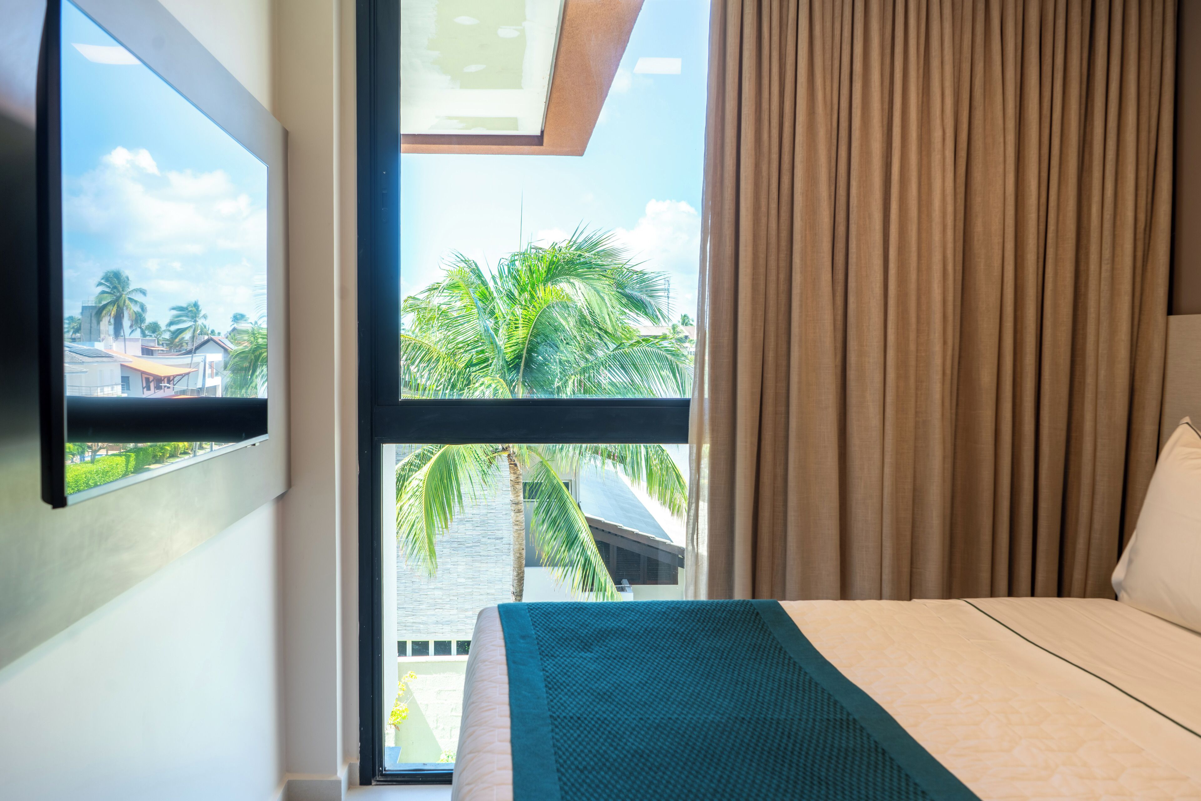 Quarto de hotel com uma janela grande, uma cama e vista para palmeiras e edifícios.