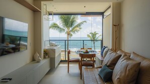 A modern living room with a large window overlooking the ocean, a sofa, a dining table with chairs, and a kitchen area. - Cais Eco Residência Muro Alto by Sintta (Ipojuca)