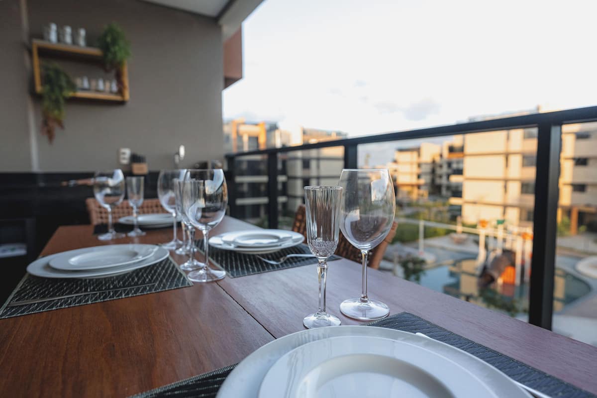 Mesa de comedor dispuesta con platos y vasos, con vistas a una piscina y edificios.
