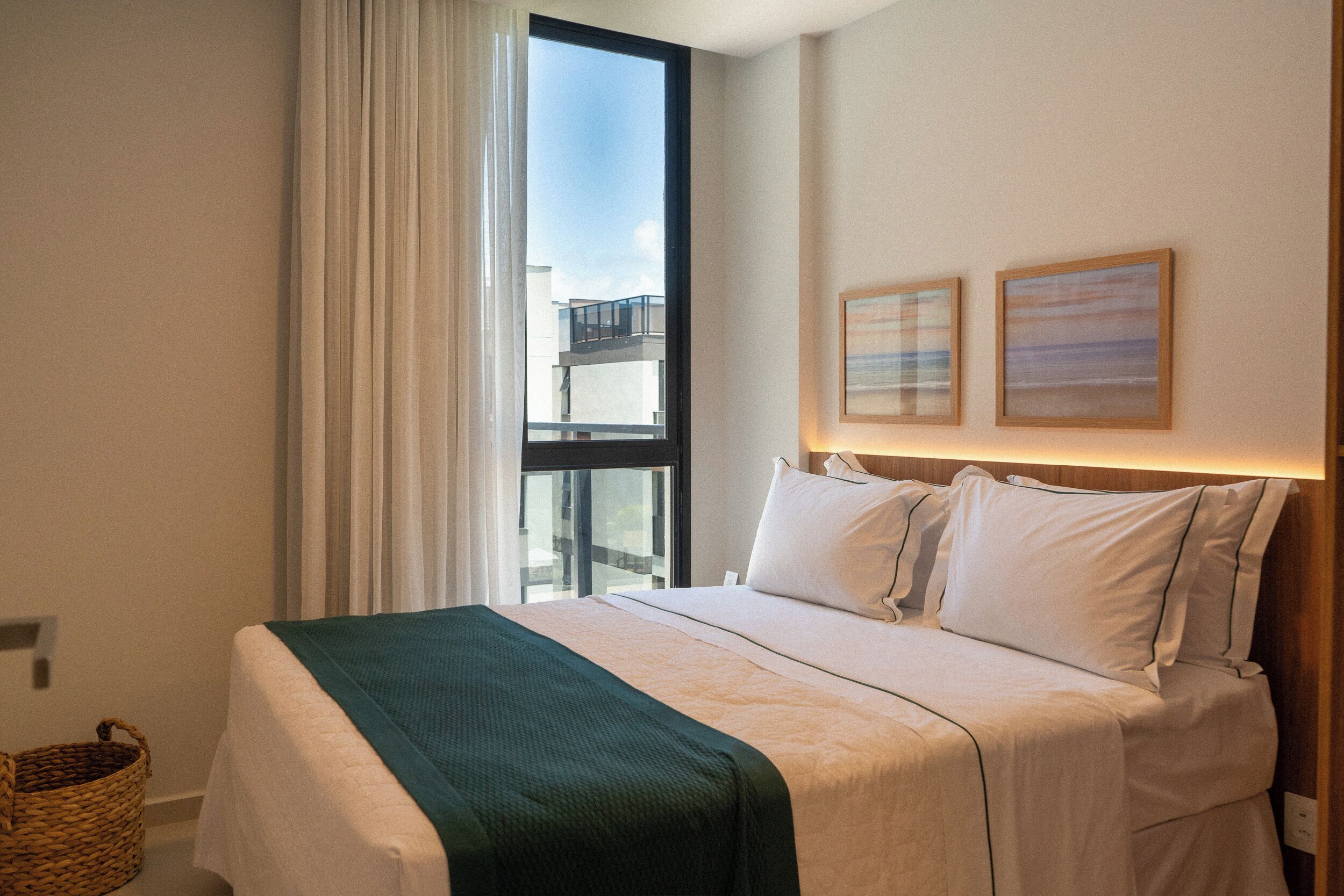 Habitació d'hotel amb una finestra gran, un llit amb roba de llit blanca i una manta verda, i dos quadres amb marc a la paret.