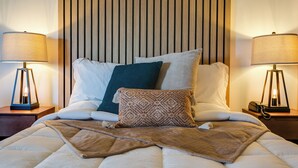 Una cama bien hecha con una manta marrón y varios cojines, flanqueada por dos mesitas de noche con lámparas.
