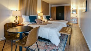 A hotel room with a large bed, two bedside tables with lamps, a chair, a small round table, a wardrobe, and a bathroom with a sink and mirror.