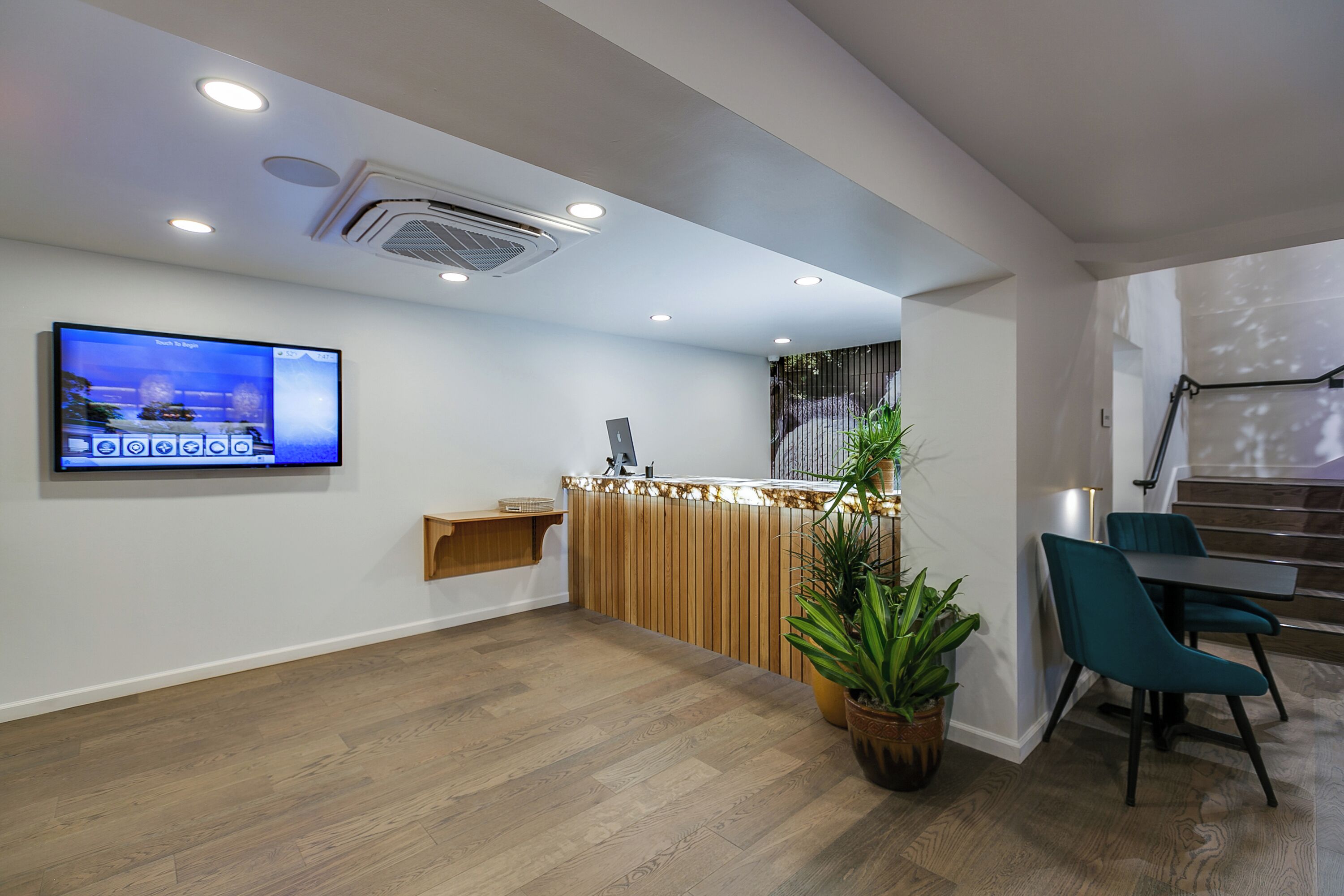 A reception area with a wooden reception desk, a mounted television, a potted plant, teal chairs, and a staircase in the background.