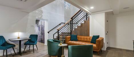 A modern living room with a staircase, a leather sofa, teal armchairs, and a small table with a lamp.