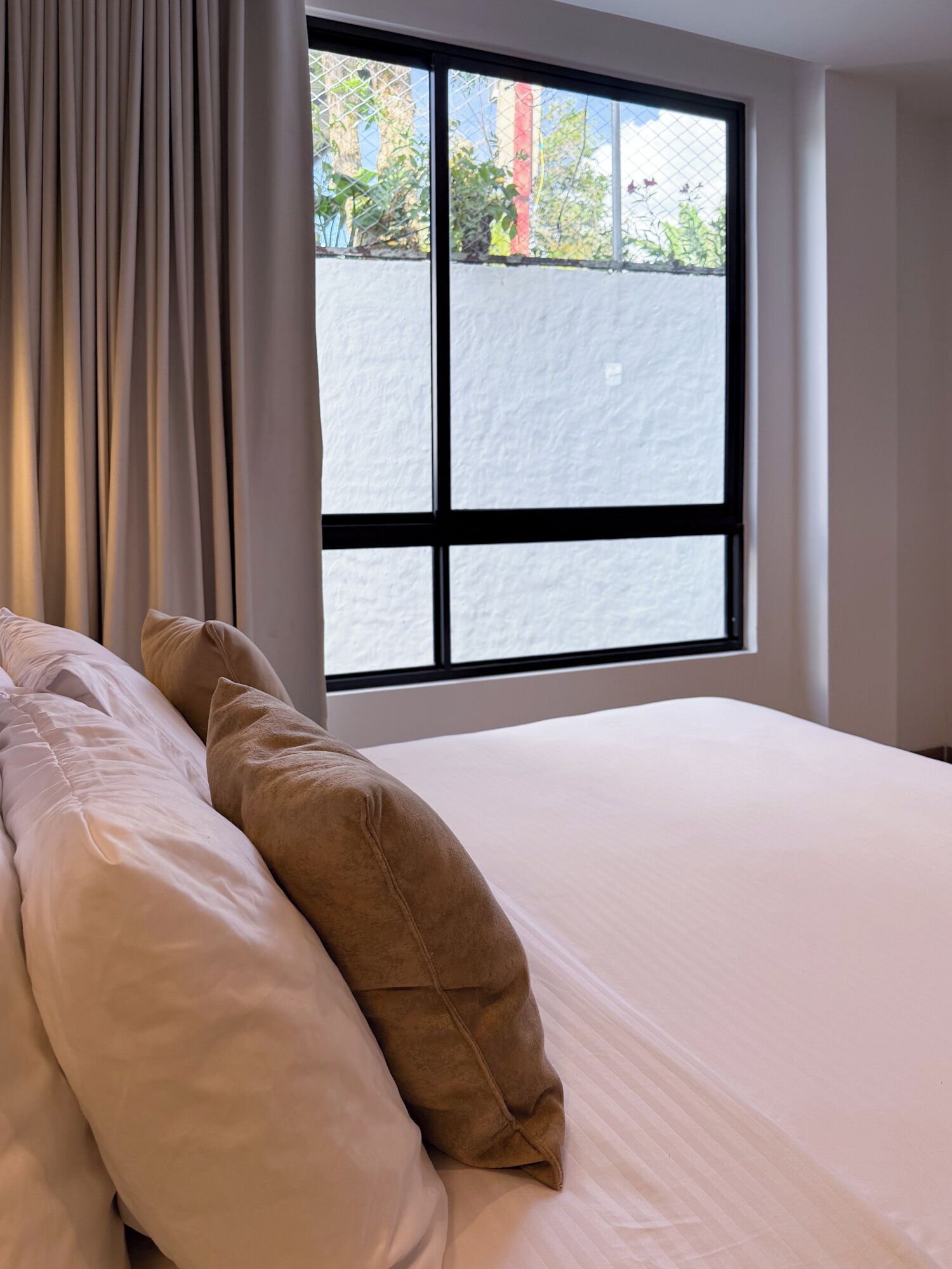 Une chambre avec une grande fenêtre, un lit avec des oreillers blancs et bruns, et une vue sur la verdure à l’extérieur.