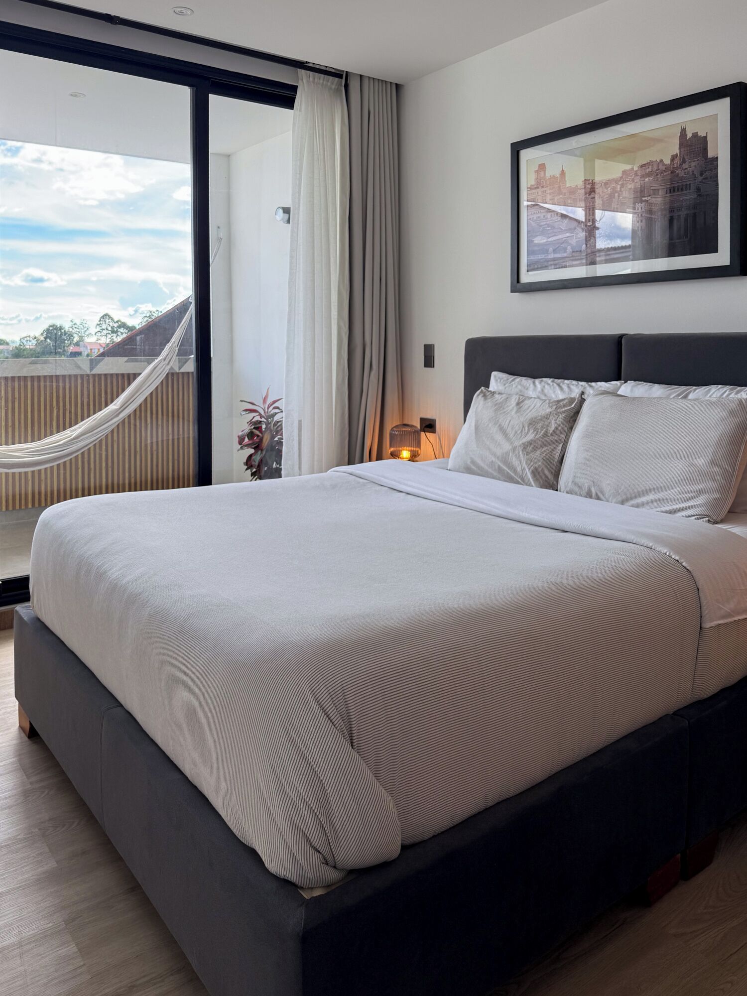 A modern hotel room with a large bed, a cityscape view, and a hammock by the window.