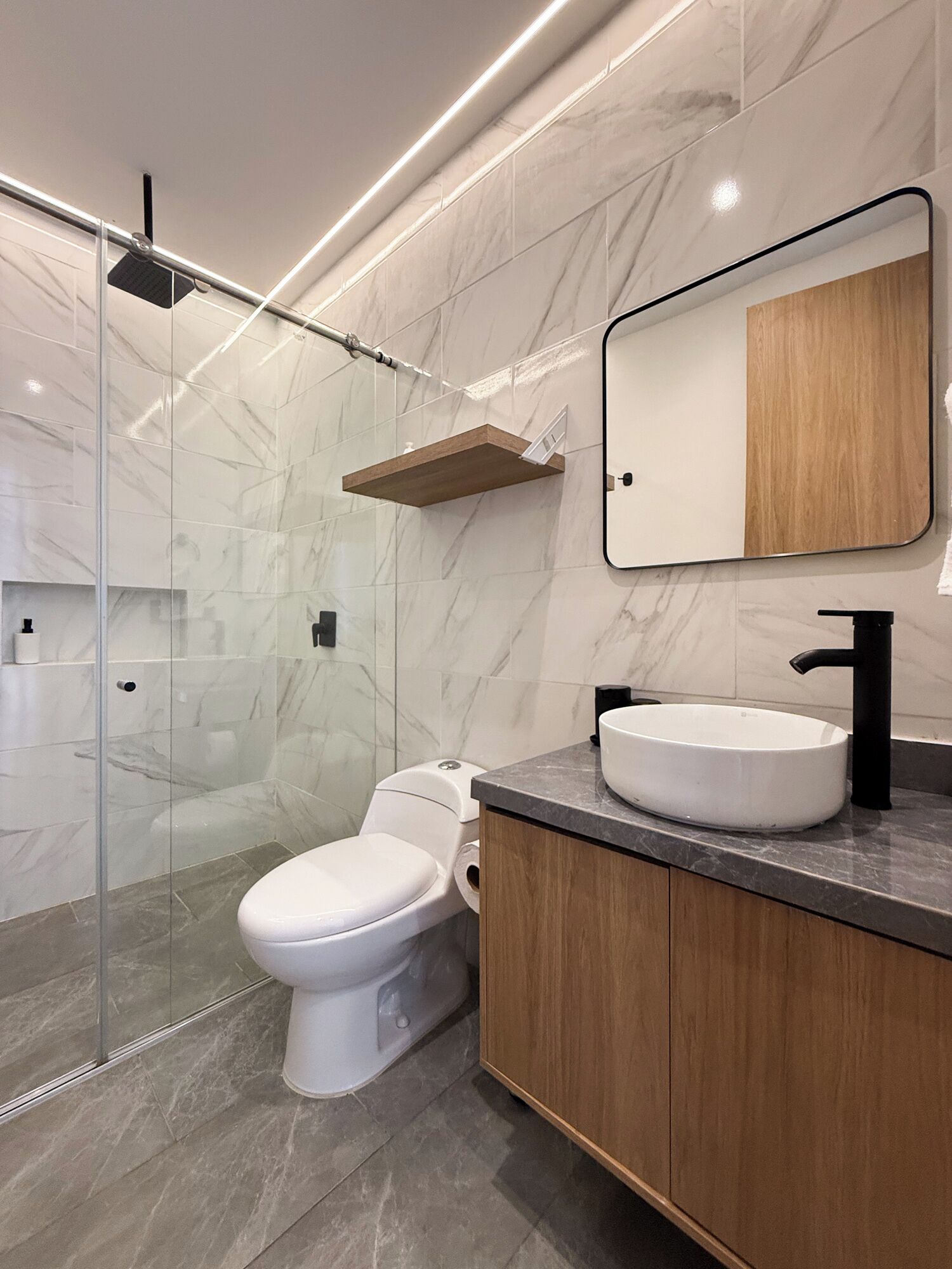 Ein modernes Badezimmer mit einer Duschkabine aus Glas, einer weißen Toilette, einem hölzernen Waschtisch mit schwarzem Waschbecken und einer Wand mit Marmorfliesen.