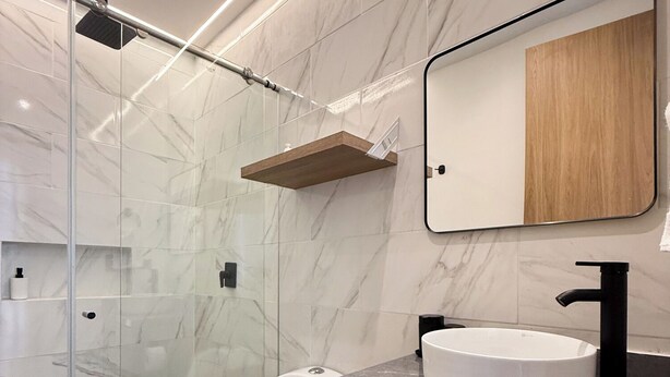 Ein modernes Badezimmer mit einer Duschkabine aus Glas, einer weißen Toilette, einem hölzernen Waschtisch mit schwarzem Waschbecken und einer Wand mit Marmorfliesen.