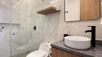 Un baño moderno con cabina de ducha de vidrio, inodoro blanco, tocador de madera con lavamanos negro y pared con azulejos de mármol.