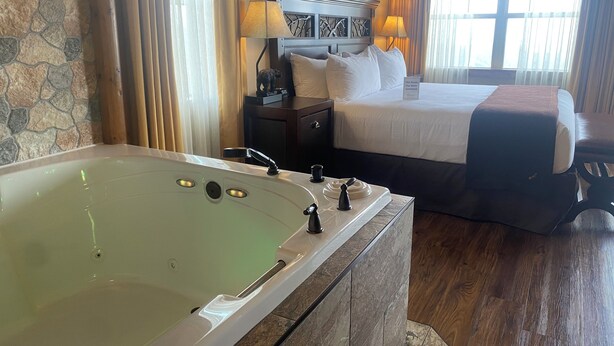 A hotel room with a large bathtub, a bed with white linens, a nightstand with lamps, and a stone wall.