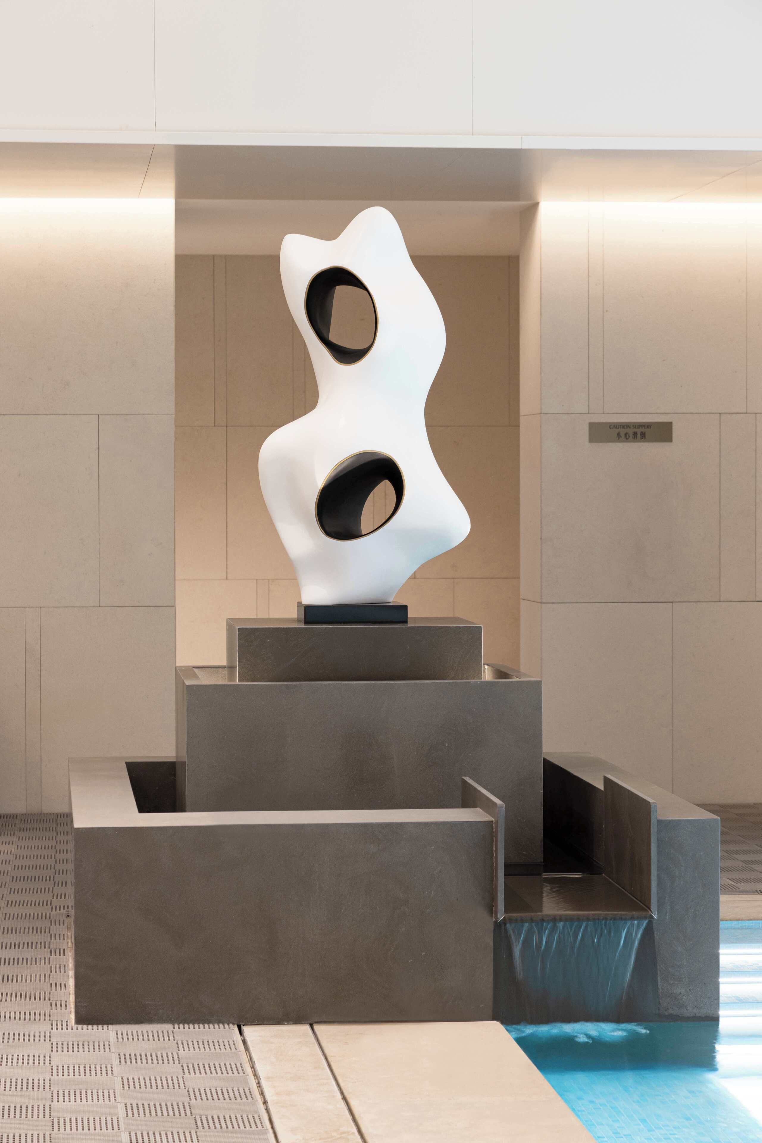 A modern sculpture with abstract shapes and cutouts, placed on a platform in a minimalist indoor setting with tiled flooring and a pool in the background.