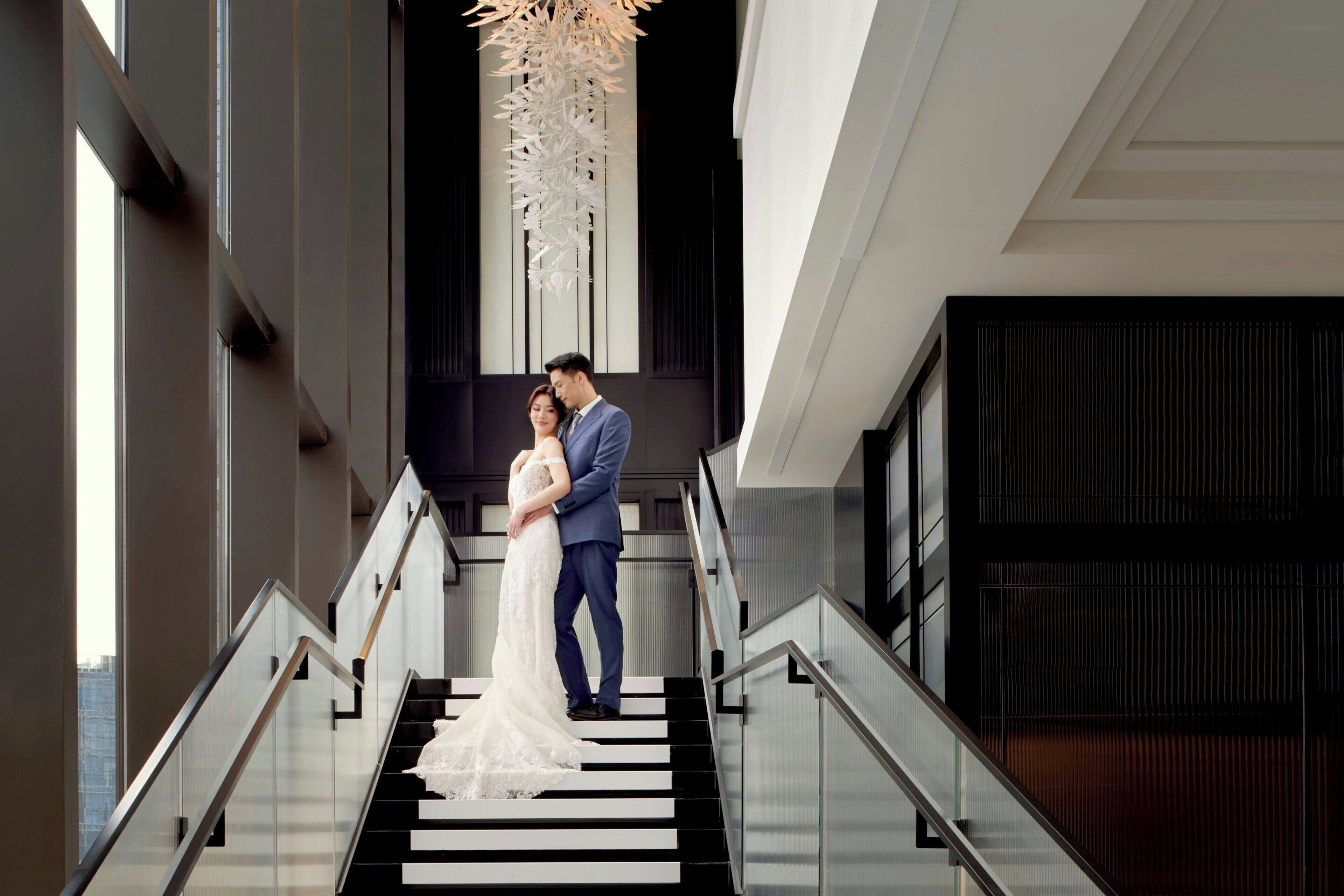 A couple on a modern staircase with glass railings, a geometric light fixture, and a view of the city.